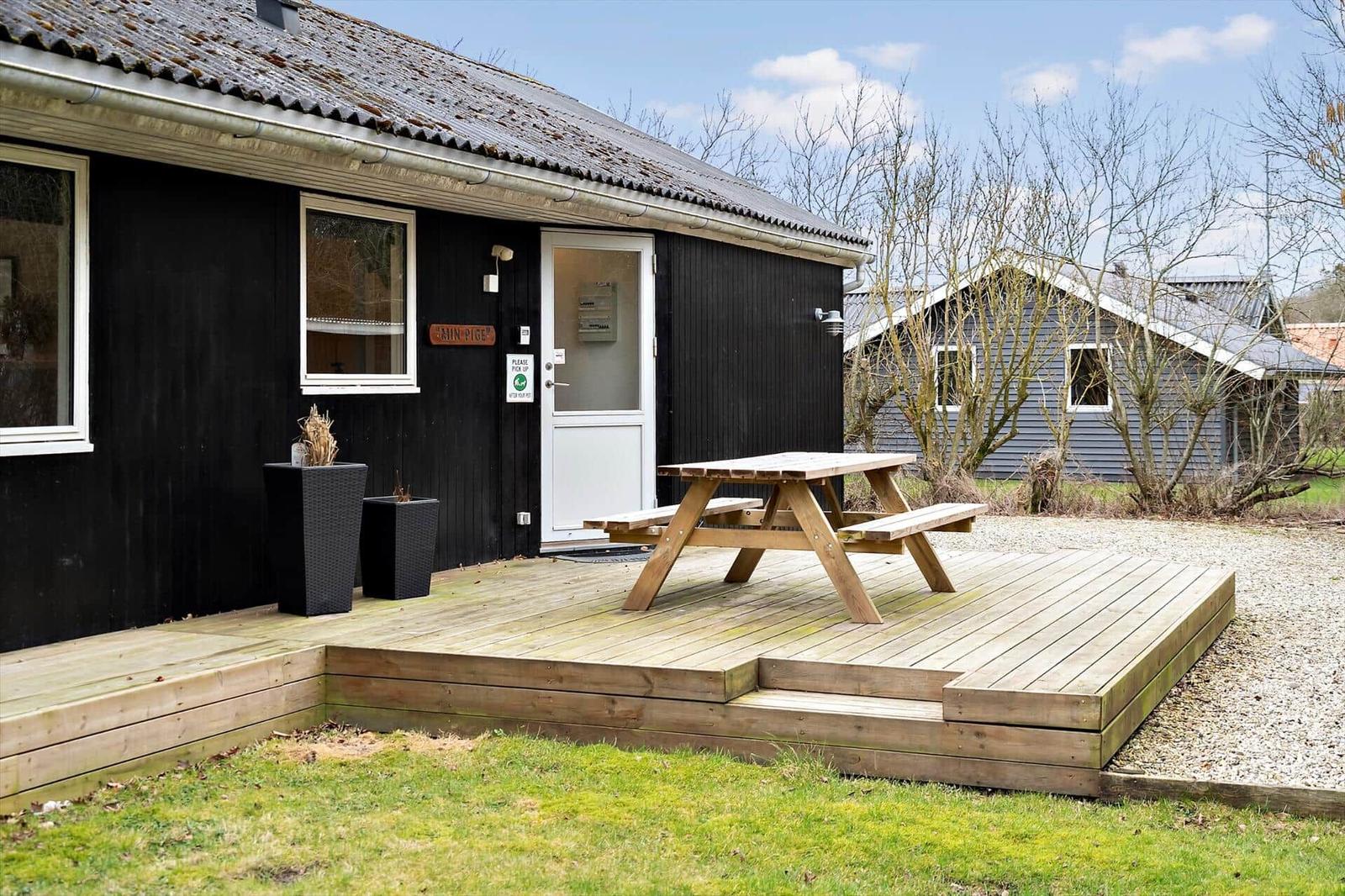 Holzterrasse mit Picknicktisch vor schwarzem Ferienhaus mit weißen Fenstern und Tür.