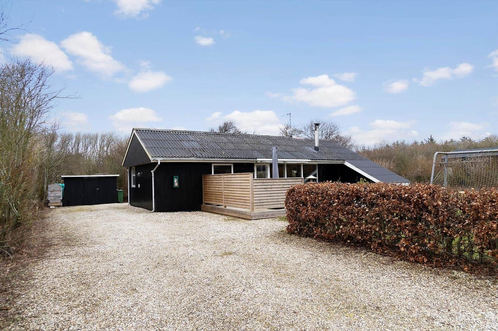 Schwarzes Ferienhaus mit Holzterrasse und Kiesfläche. Hinterhof mit Hecke und Tor.