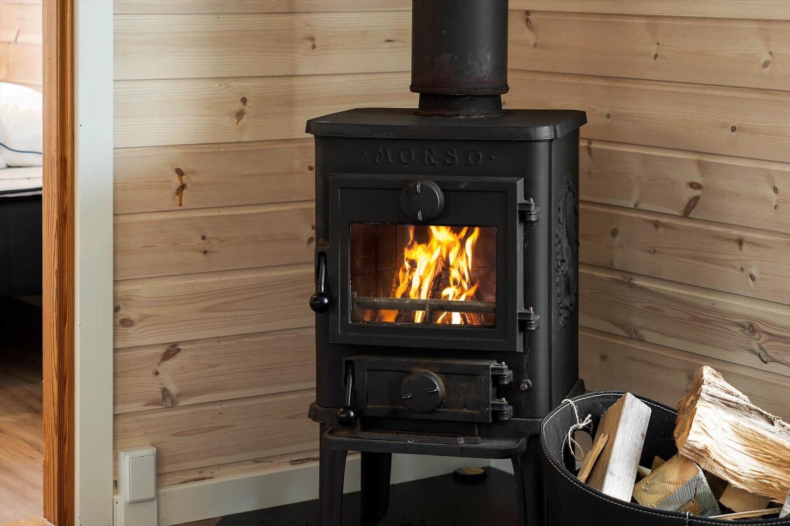 Holzofen mit brennendem Feuer in einem Raum mit Holzwandverkleidung und Holzstapel daneben.