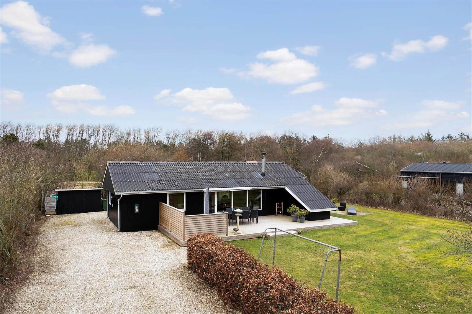 Schwarzes Haus mit Terrasse und Garten. Hintergrund Wald. Himmel blau mit Wolken.