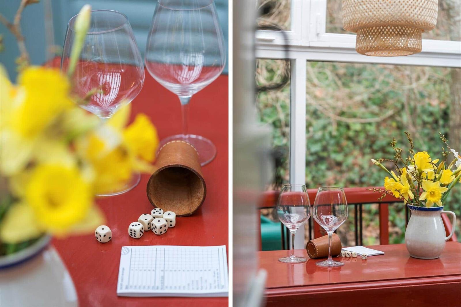 Roter Tisch mit Weinflaschen, Würfeln und Blumen vor Fenster.