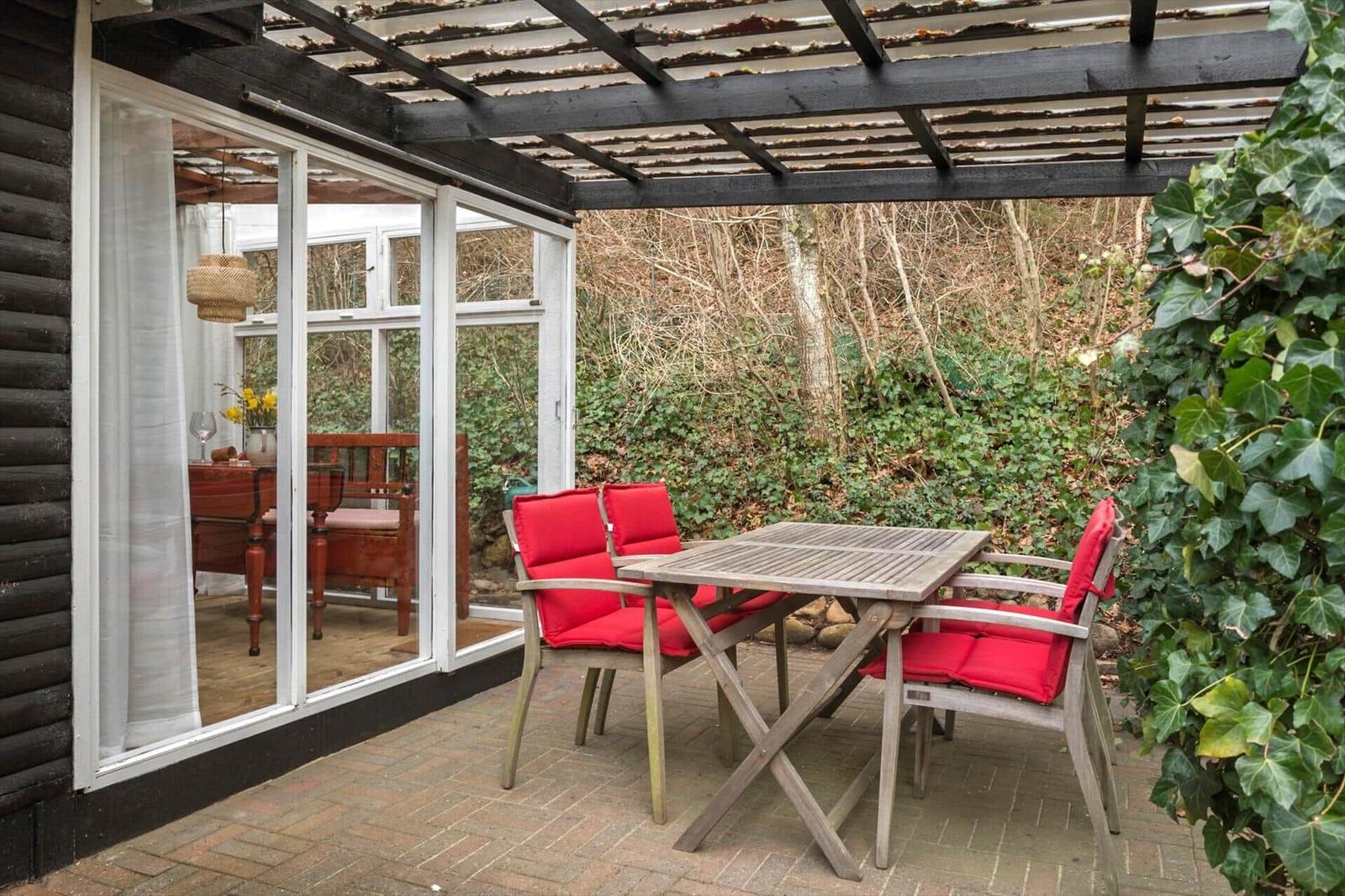 Terrasse mit Tisch und roten Sitzkissen unter Holzüberdachung. Blick in das Haus mit Essbereich.