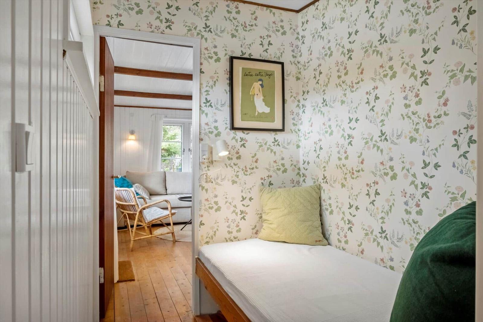 Schlafzimmer mit floraler Tapete und Bett. Durchgang in Wohnzimmer mit Sofa und Stuhl.