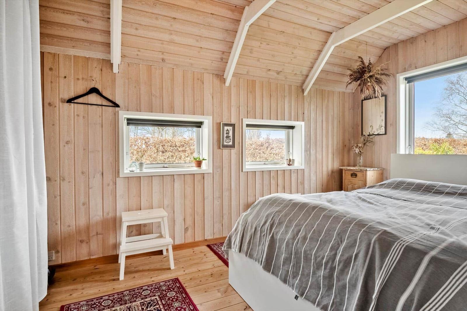 Schlafzimmer mit Holzwand, Bett, Fenstern und Stufenstuhl.