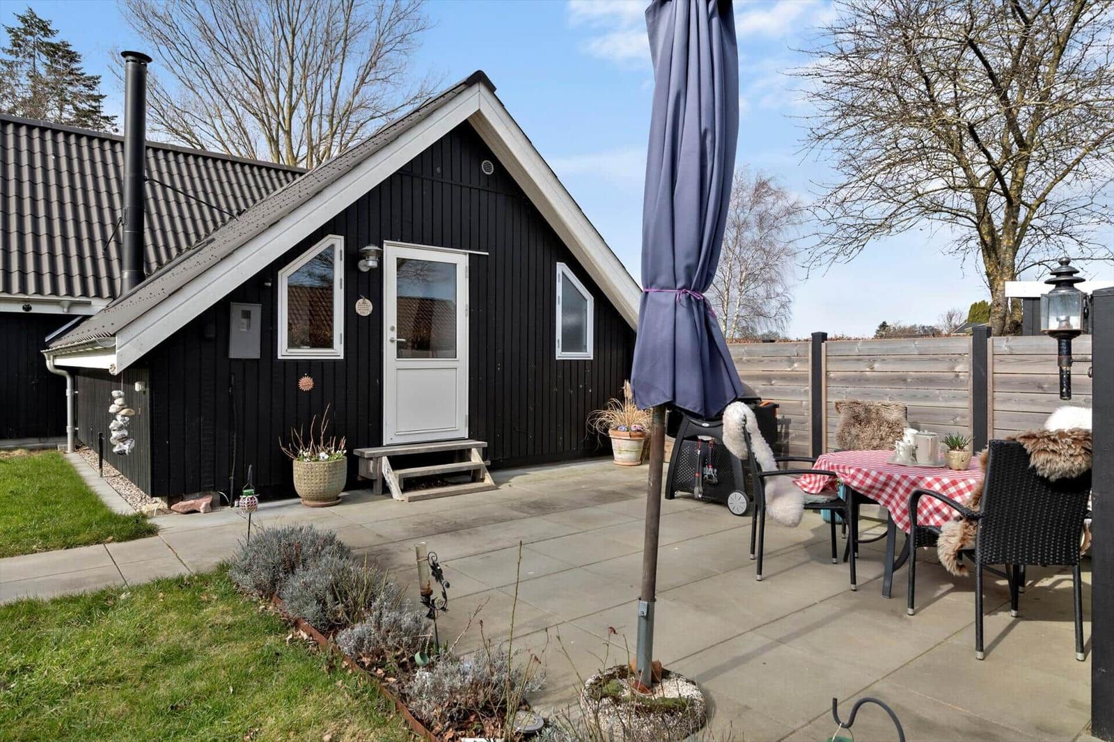 Schwarzes Holzhaus mit Terrasse und Garten. Tisch mit rot-weißem Tischtuch und Stühlen.