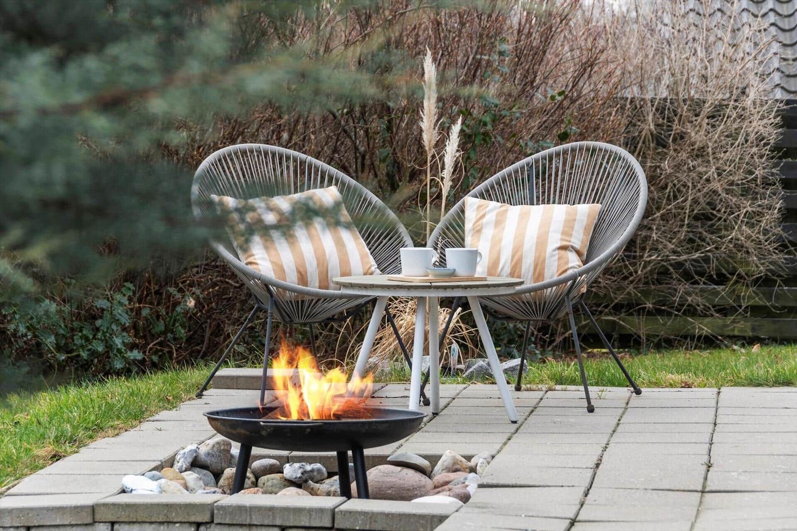 Zwei Sessel mit Kissen und Tisch stehen neben einem brennenden Feuerkessel im Garten.