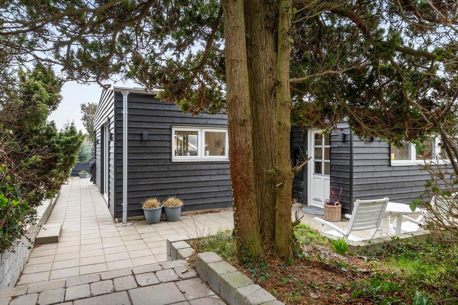 Ein graues Holzhaus mit weißer Tür und Fenstern. Außenbereich mit Sitzgelegenheit und Bäumen.