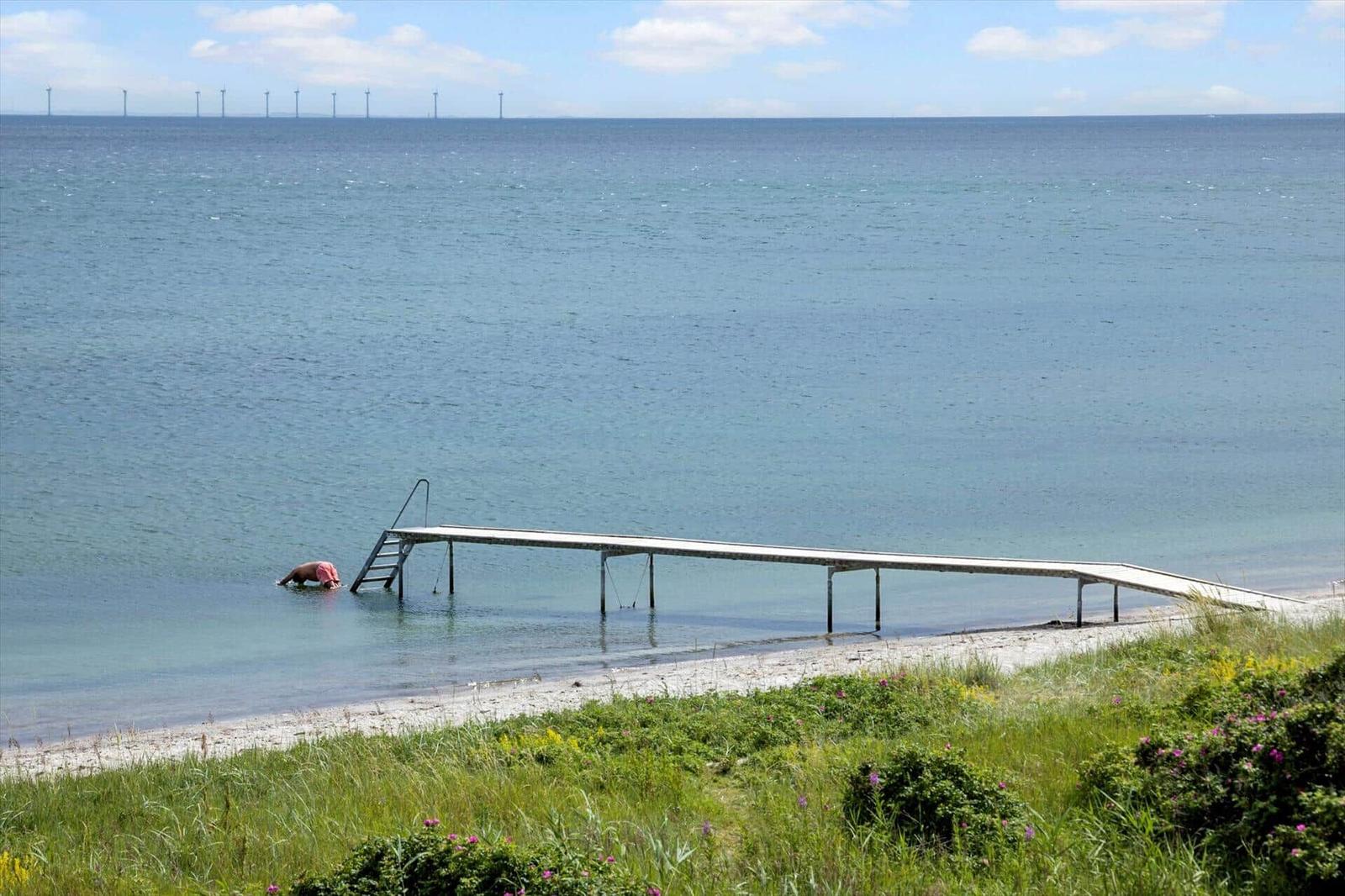 Ein Steg führt ins Meer. Eine Person taucht im Wasser. Windkraftanlagen sind im Hintergrund sichtbar.