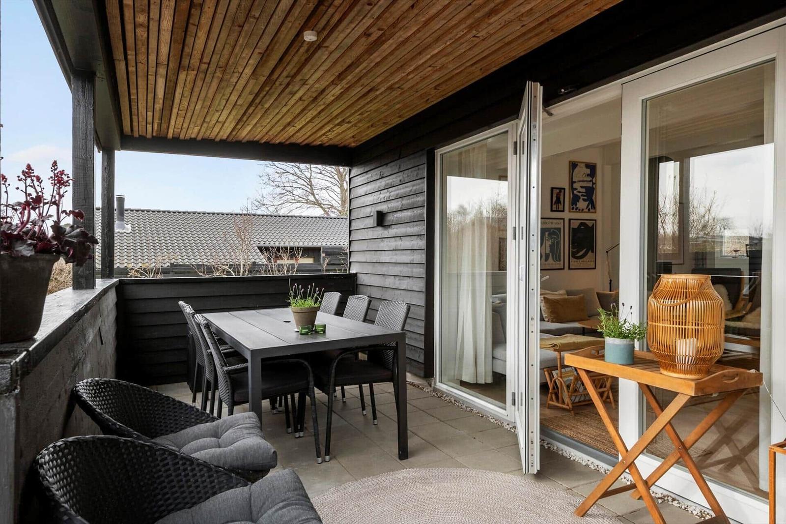 Terrasse mit Tisch, Stühlen und Pflanzen. Durchgehende Glastüren führen ins Innere.