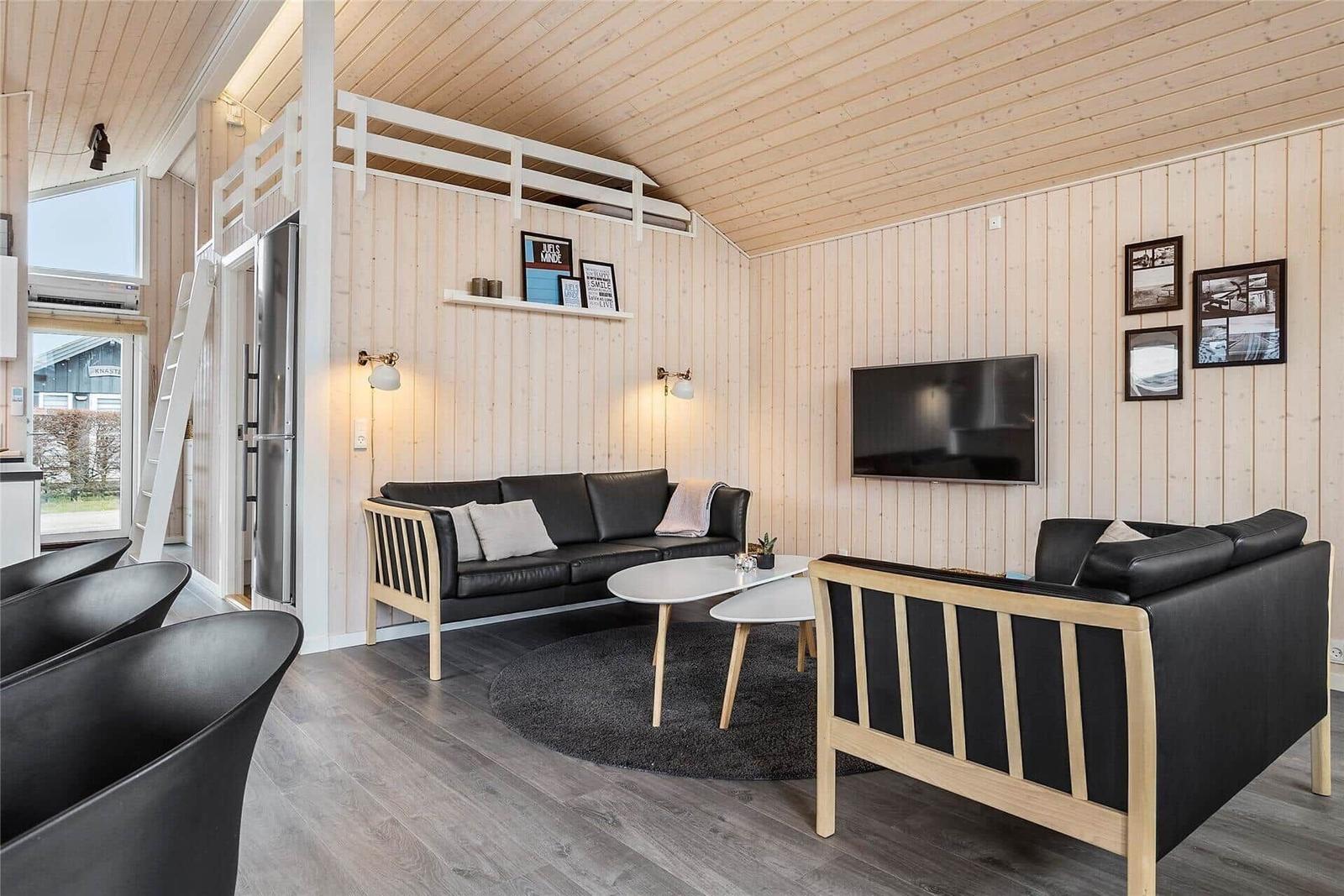 Wohnzimmer mit Sofa, Sessel und Fernseher an Holzwand. Obergeschoss mit Schlafplatz.