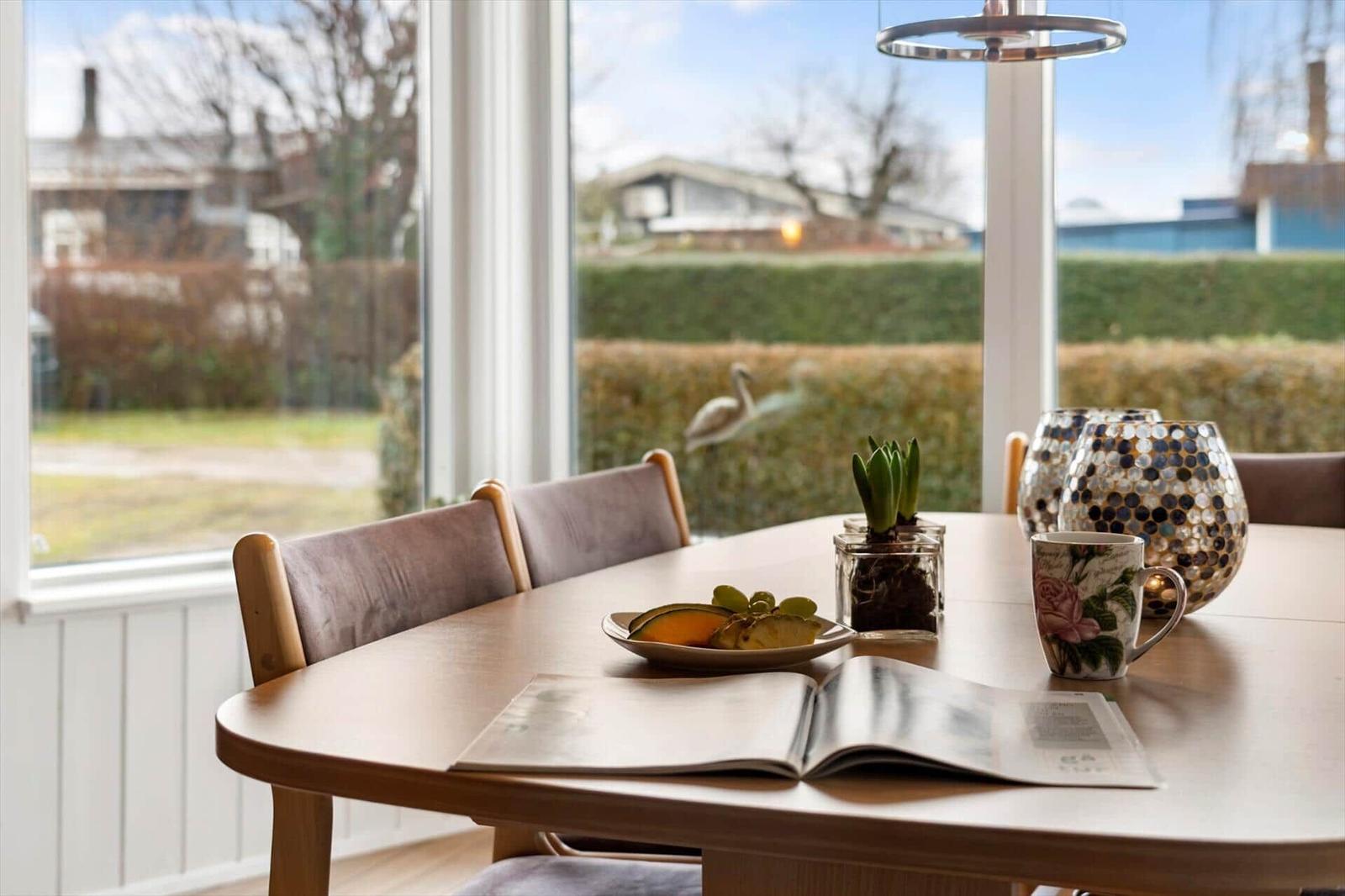 Esstisch mit Stühlen, Tasse, Buch und Pflanzen vor Fenster mit Blick auf Garten.