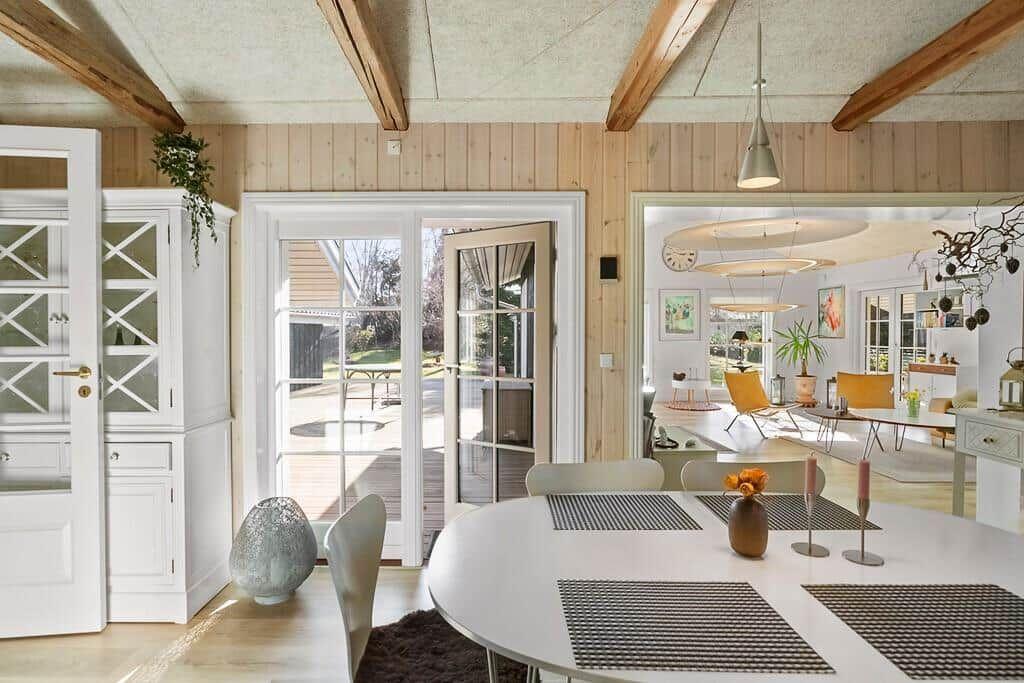 Dining area with round table, white chairs, and view to garden. Wooden ceiling and white furniture.