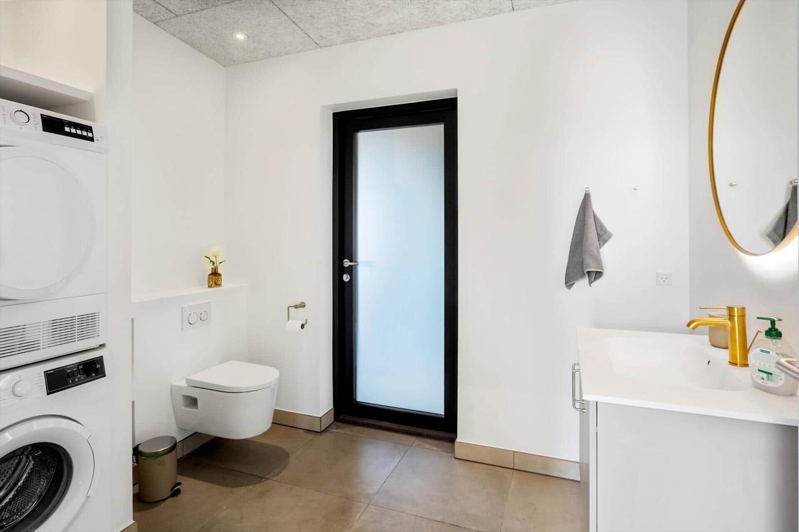 Bathroom with washing machine, toilet, and sink with gold faucet.