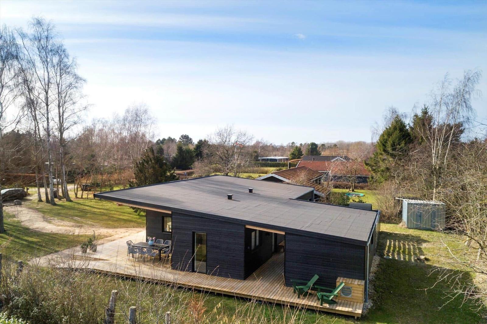Modern villa with wooden terrace and garden. Greenery and trees surround the house.