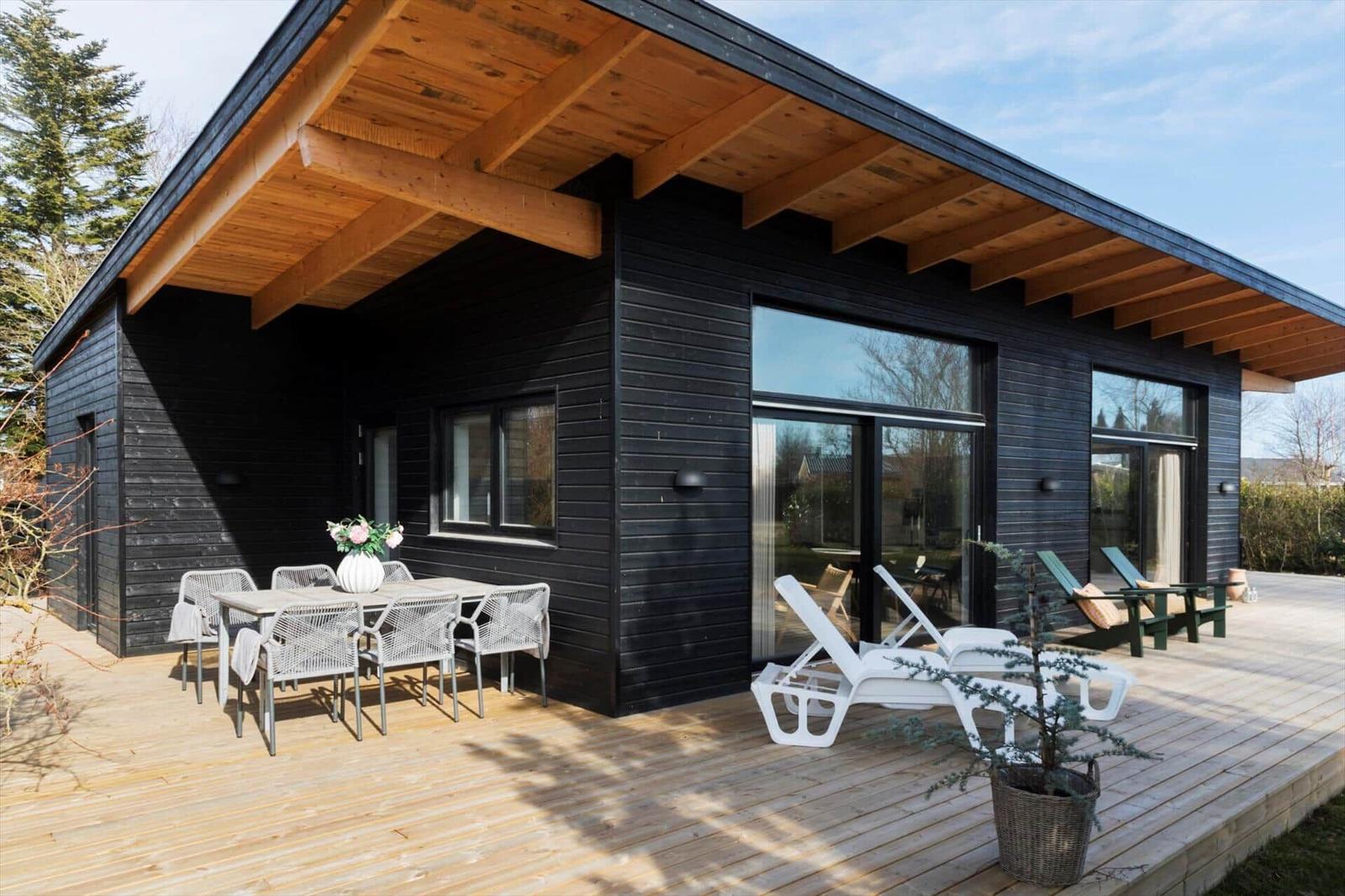 Modern villa with wooden terrace, seating group, and lounge chairs. Black cladding and large windows.