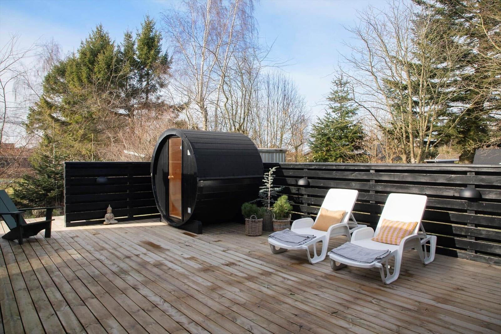 Deck with sauna, lounge chairs, and wooden railing. Background: trees and clear sky.