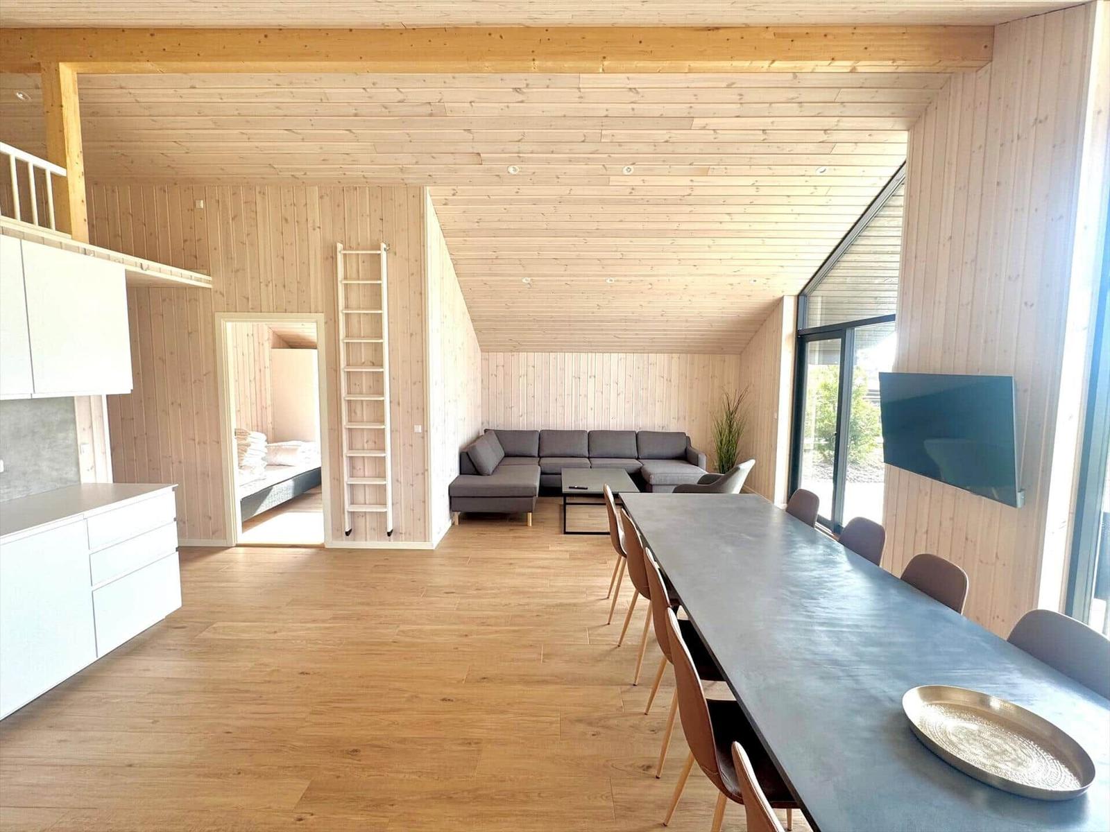 Living area with dining table, sofa, and kitchen. Wooden ceiling and walls. TV and bedroom in background.