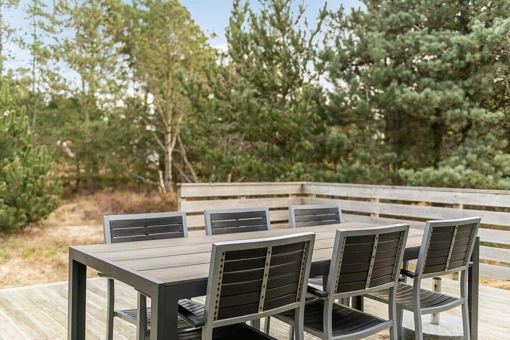 Holzterrasse mit Tisch und sechs Stühlen, umgeben von Bäumen.