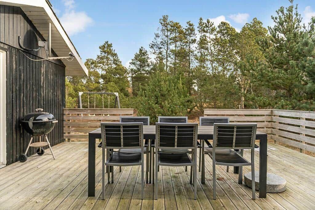 Terrasse mit Tisch, Stühlen und Grill. Hintergrund: Wald und Himmel.