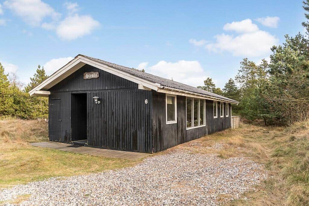 Schwarzes Holzhaus mit Dach, Fenstern und Eingang. Umgeben von Bäumen und Kiesweg.