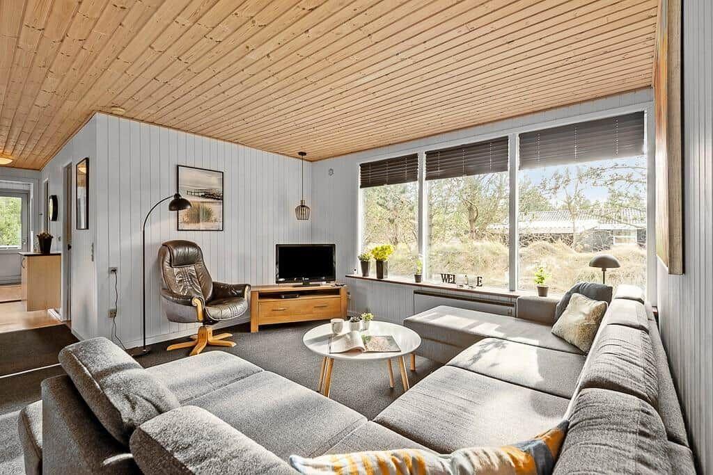 Wohnzimmer mit Sofa, Sessel und TV. Große Fenster mit Blick nach draußen.