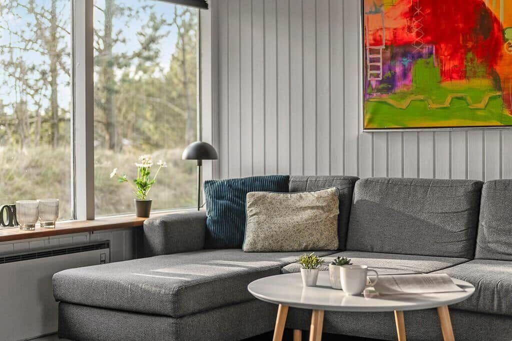 Grauer Sofa mit Kissen, runder Tisch und Fensterblick auf Wald.