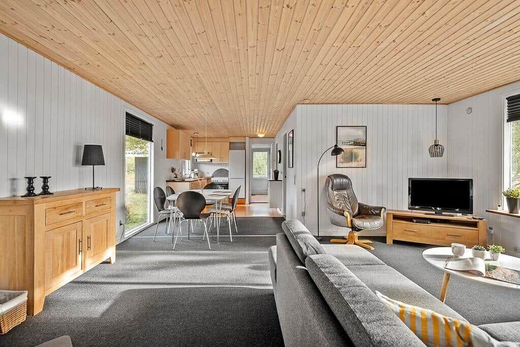 Wohnbereich mit Sofa, Sessel und TV. Küche im Hintergrund. Holzdecke und weiße Wände.