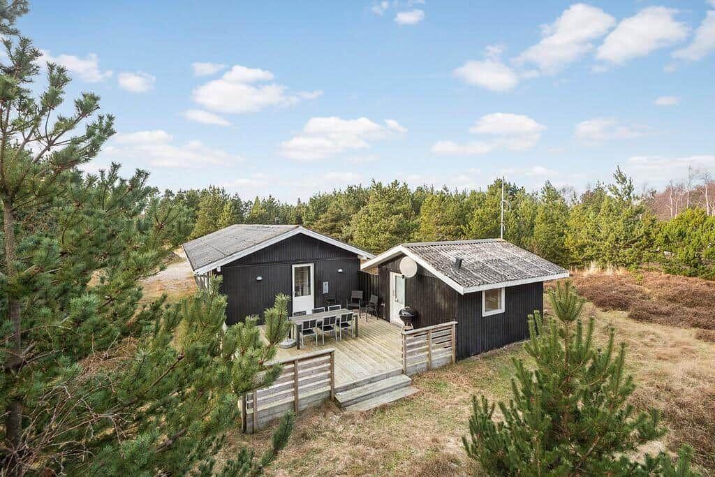 Zwei Holzhäuser mit Terrasse und Grill in Waldnähe unter blauem Himmel.