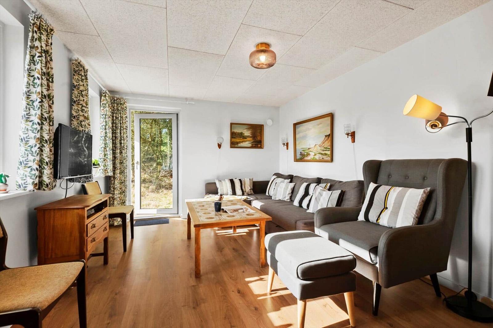Wohnzimmer mit Sofa, Sessel, Couchtisch und Fernseher. Holzboden und Fenster mit Blick auf Garten.