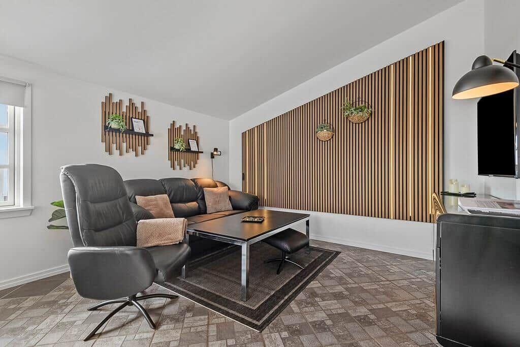Wohnzimmer mit Sofa, Sessel und Holzwand. Fenster, Teppich und Couchtisch sichtbar.
