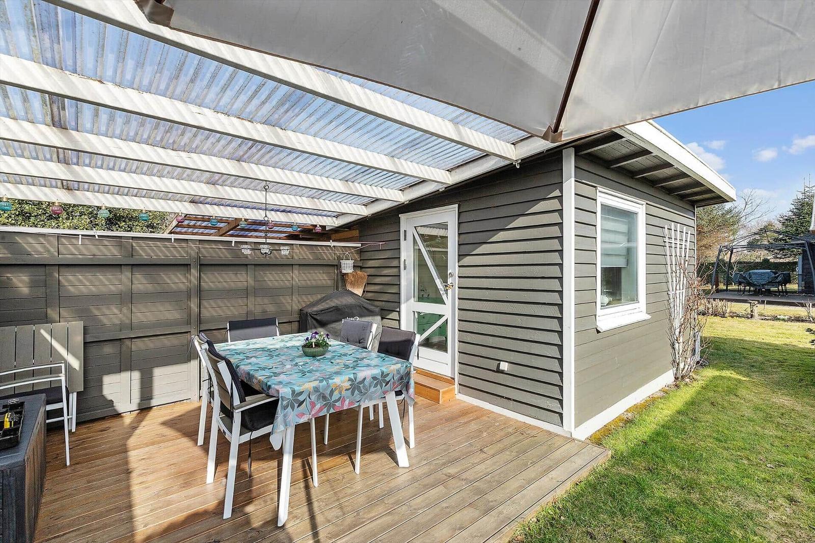 Terrasse mit Tisch, Stühlen und Sonnenschutz neben grauem Haus mit Garten.