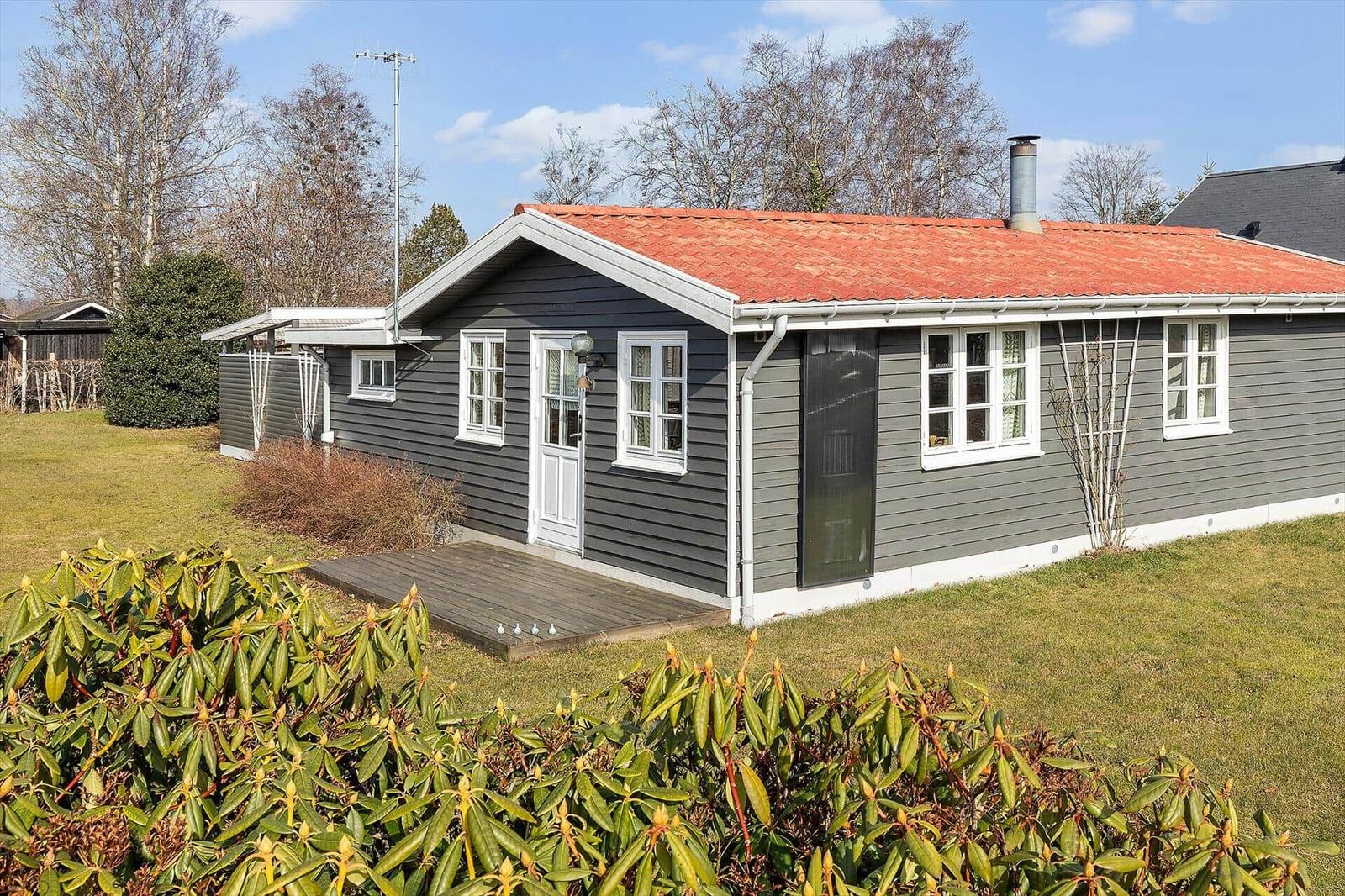 Ein graues Holzhaus mit rotem Dach und weiß gerahmten Fenstern. Vor dem Haus befindet sich eine kleine Terrasse.
