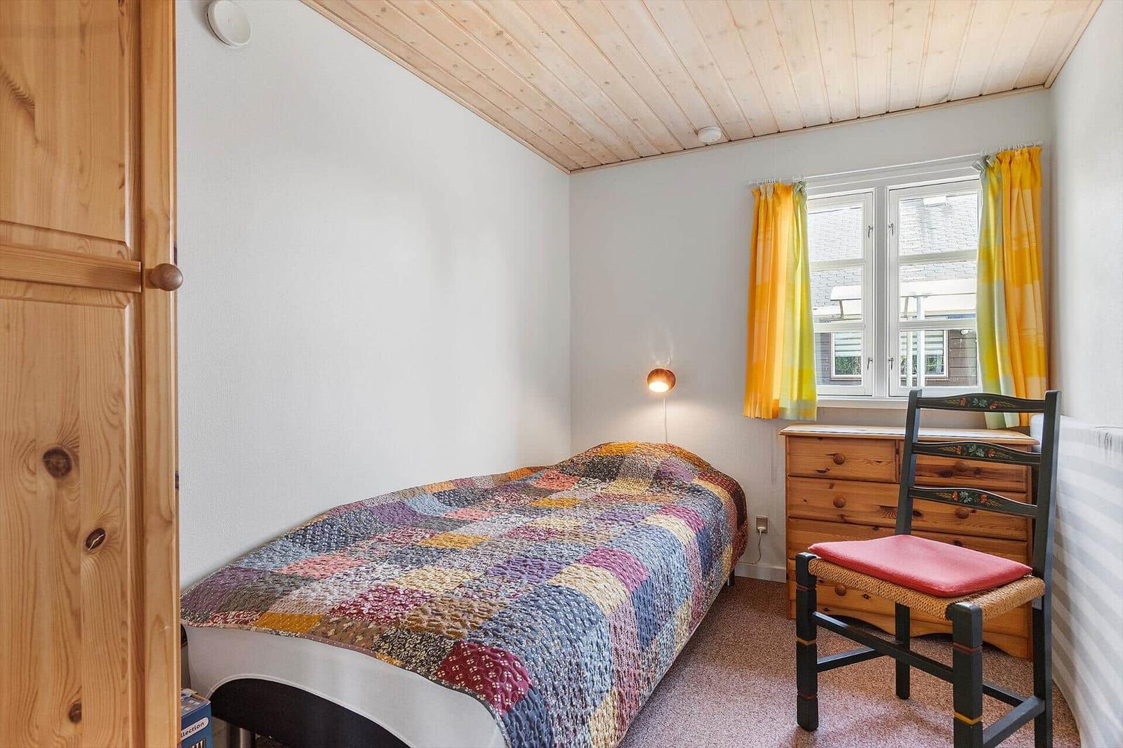 Ein Schlafzimmer mit Bett, Schrank, Stuhl und Fenster mit gelben Vorhängen.