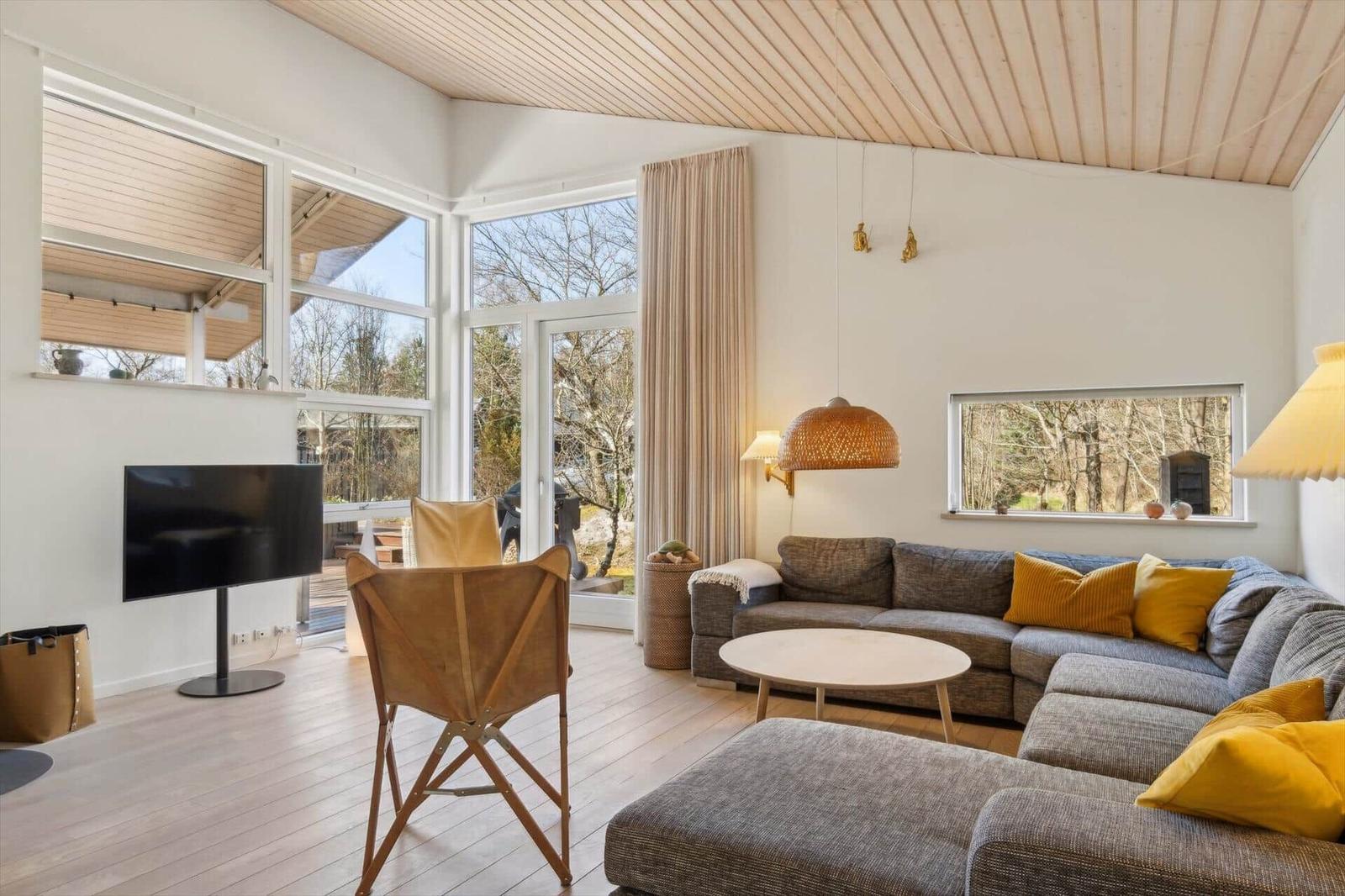 Wohnzimmer mit Sofa, Fernseher und großen Fenstern. Holzdecke und helle Wände.