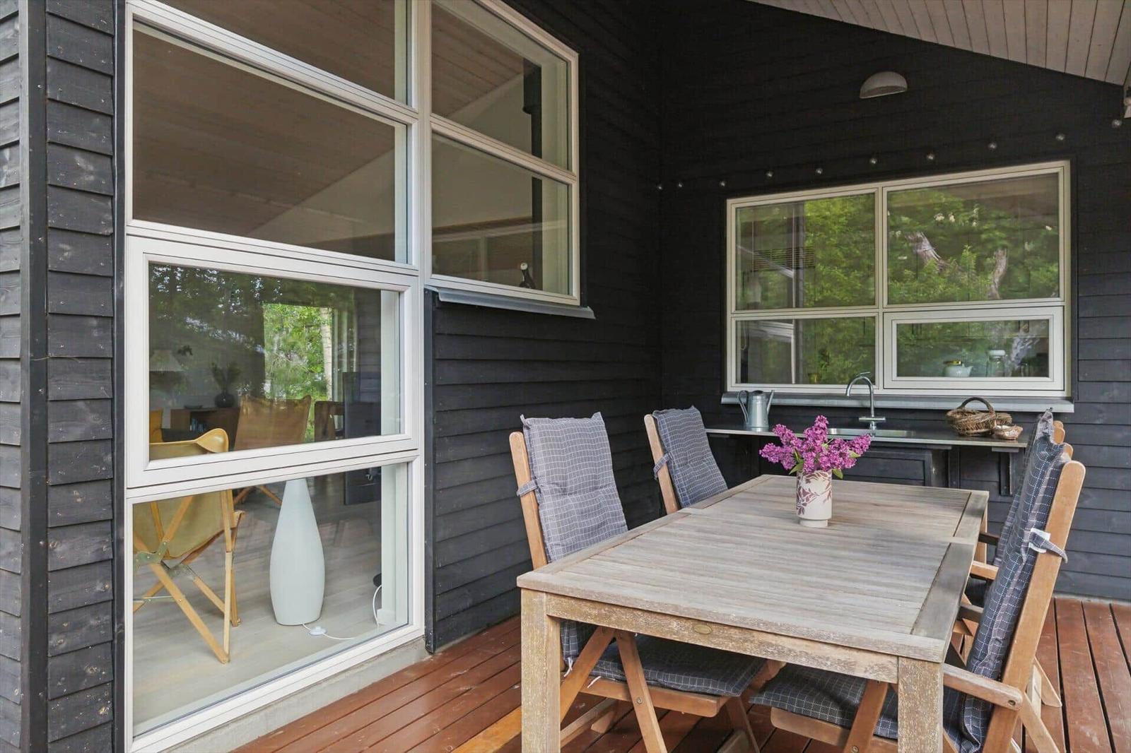 Terrasse mit Holztisch, Stühlen und Blumen. Fenster zeigen den Innenraum.