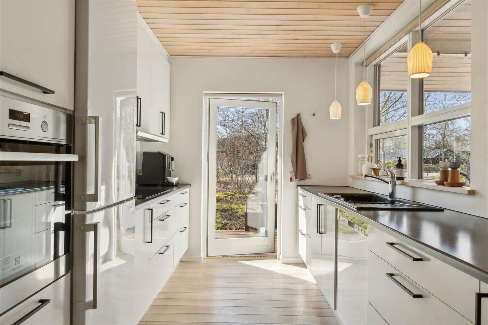Moderne Küche mit weißen Schränken, Edelstahlgeräten und Holzboden. Durchgang zur Terrasse.