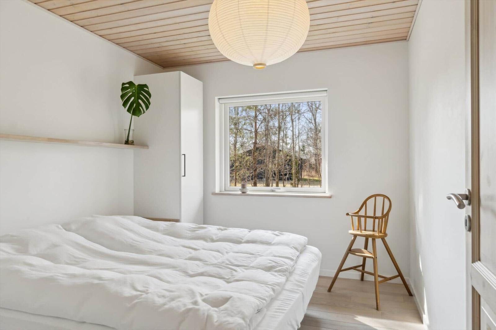 Schlafzimmer mit weißem Bett, Holzdecke und Fensterblick auf Bäume.