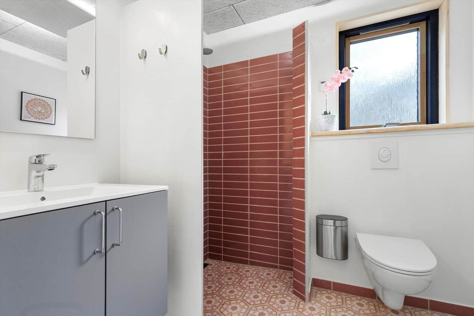 Bathroom with shower, sink, and toilet. Red tiles and window with flower.
