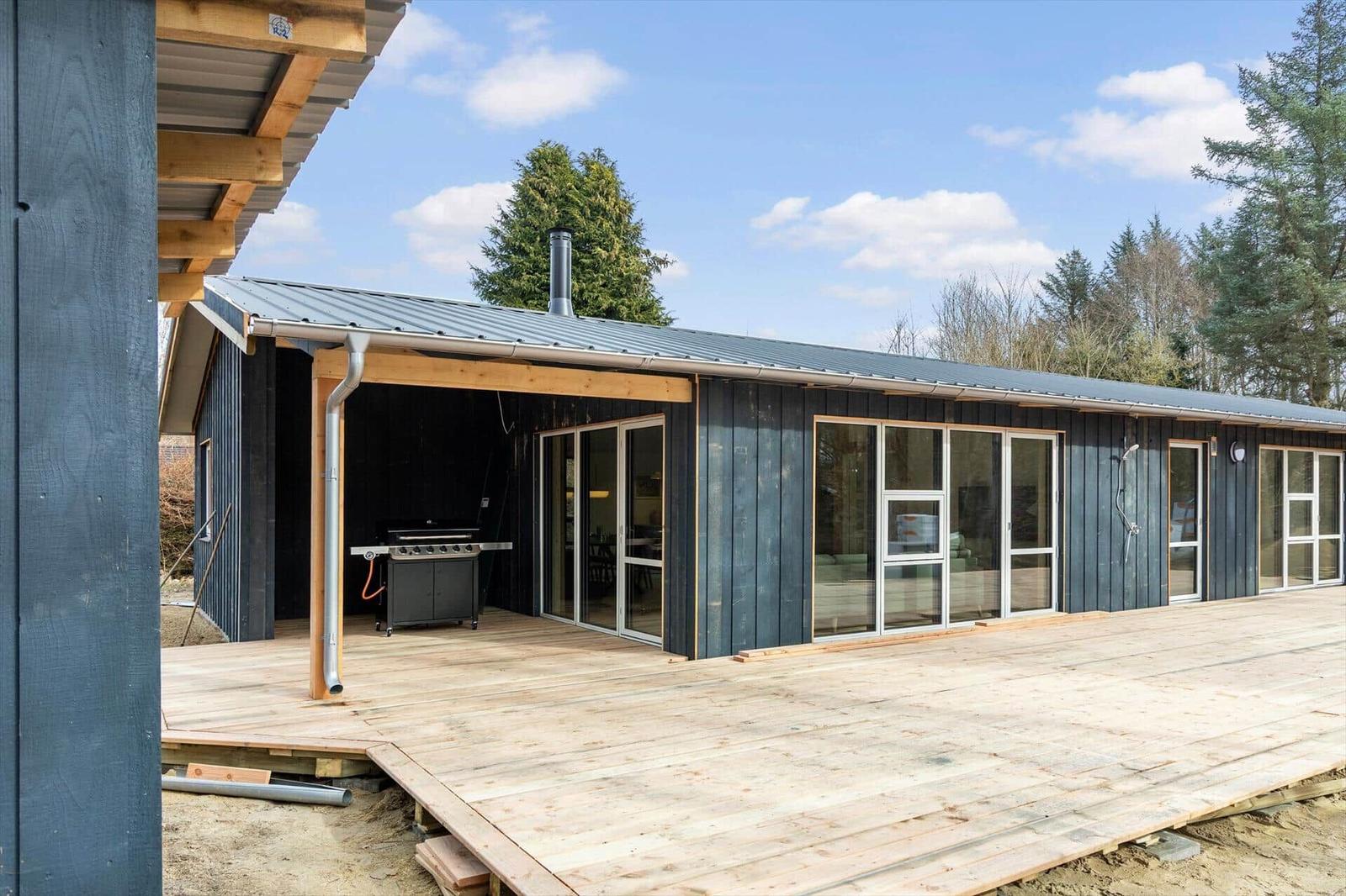 Modern wooden cabin with terrace and grill area. Large window front. Surrounded by trees.
