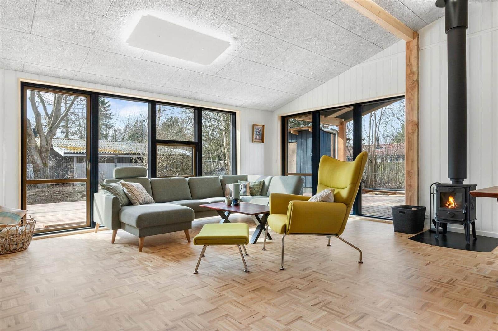 Living room with sofa, armchair, and wood stove. Parquet floor and large windows.