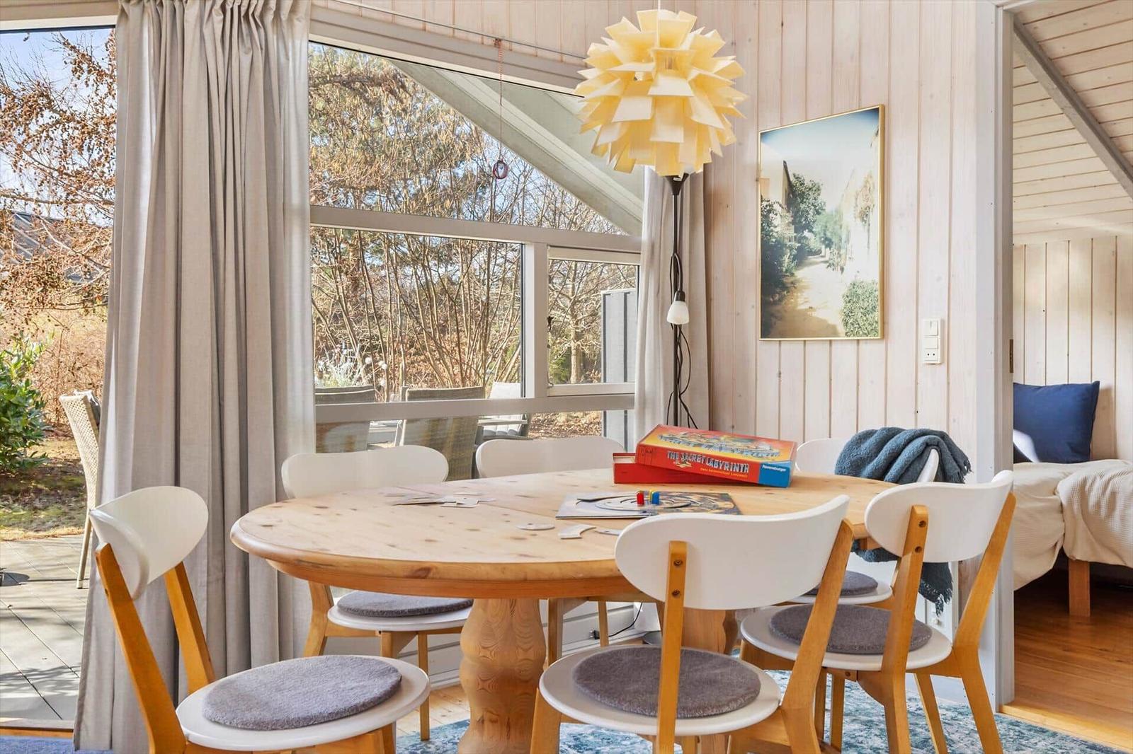 Round wooden table with chairs, garden view through window, lamp and picture on wall.