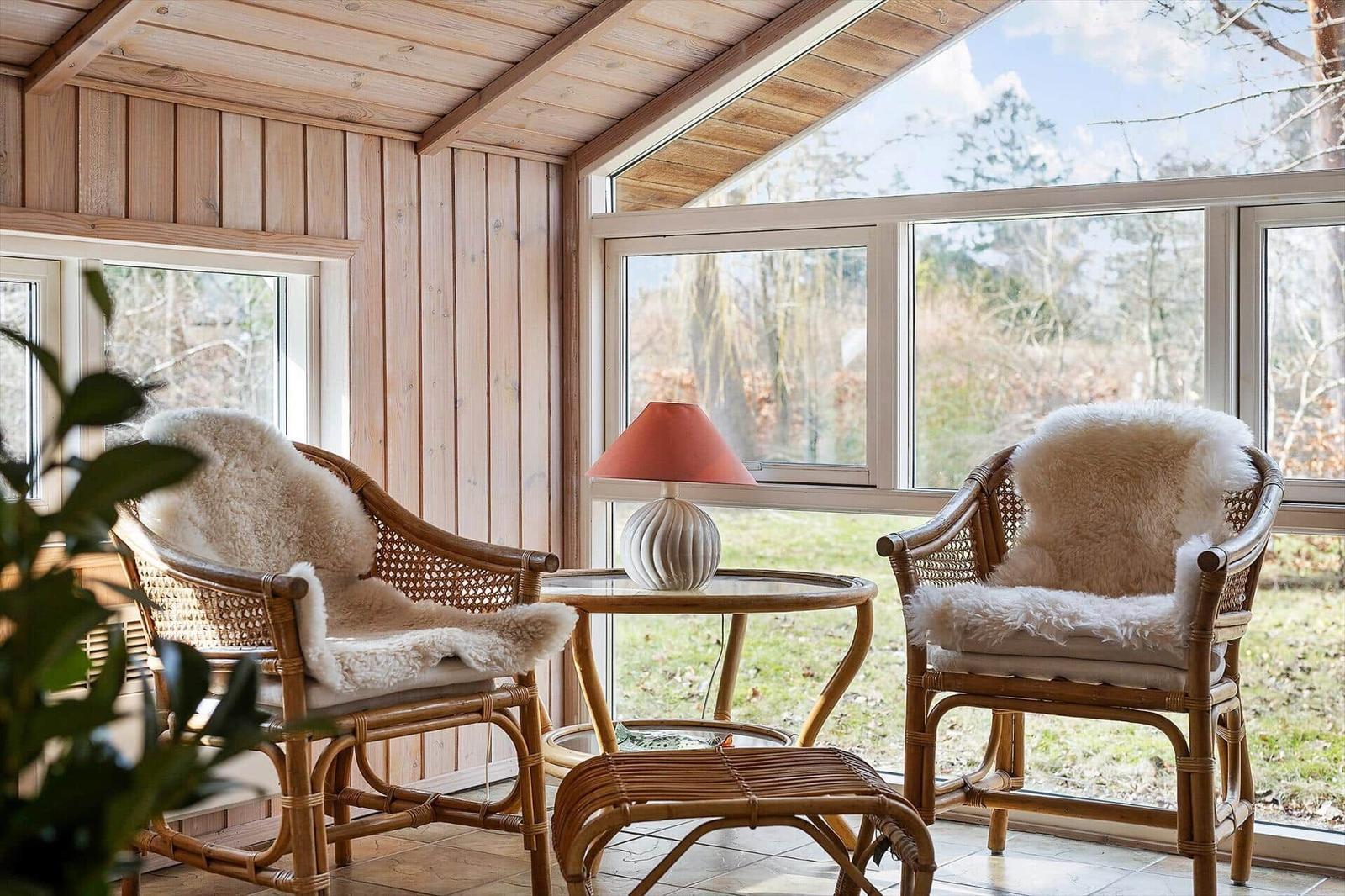Two rattan chairs with fur covers stand by large windows. Between them is a small table with a lamp.