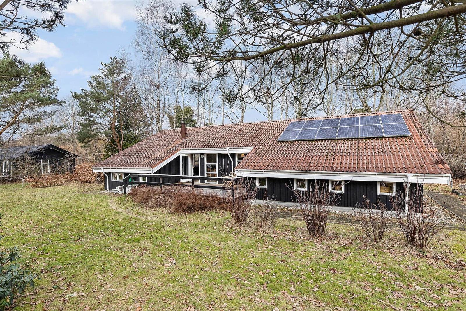 Black wooden house with solar panels on roof, surrounded by grass and trees.
