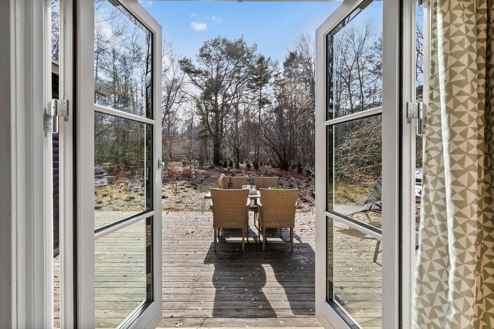 Through open doors, you see a terrace with table and chairs in the woods.