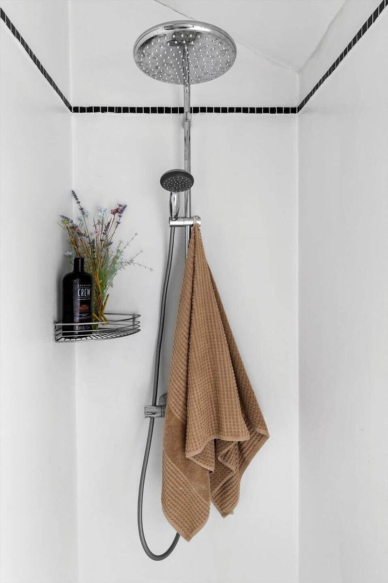 Shower stall with overhead and handheld showerheads. Towel hanging. Shelf holds bottle and dried flowers.