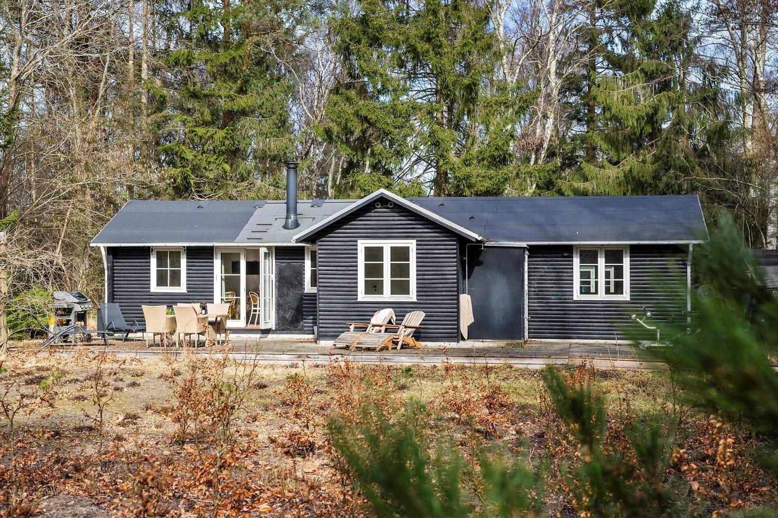 Black wooden house with terrace, garden, and forest in the background.