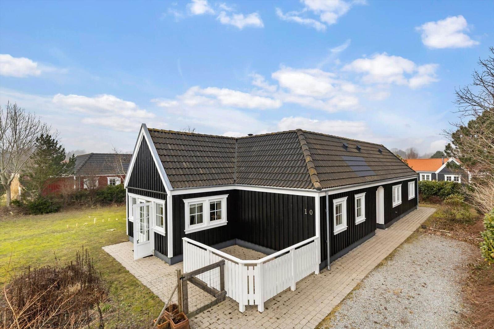 Black wooden house with white railing and tiled roof. Garden and path are visible.