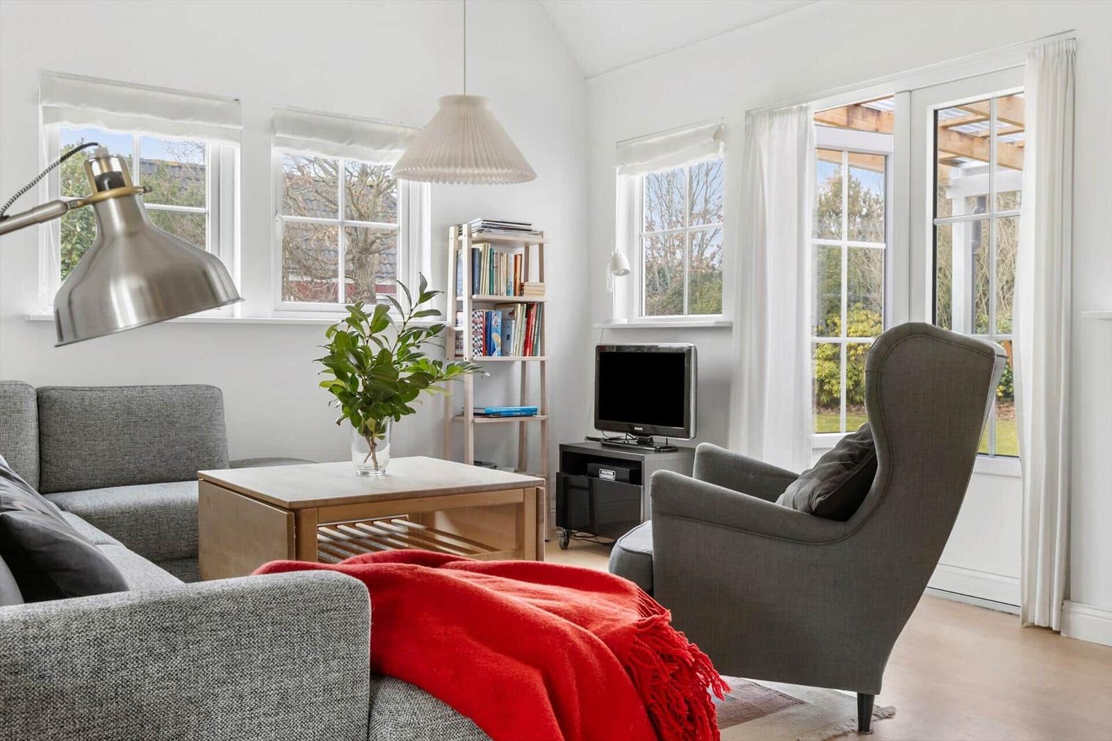 Living room with sofa, armchair, coffee table, and TV. Large windows and white walls.