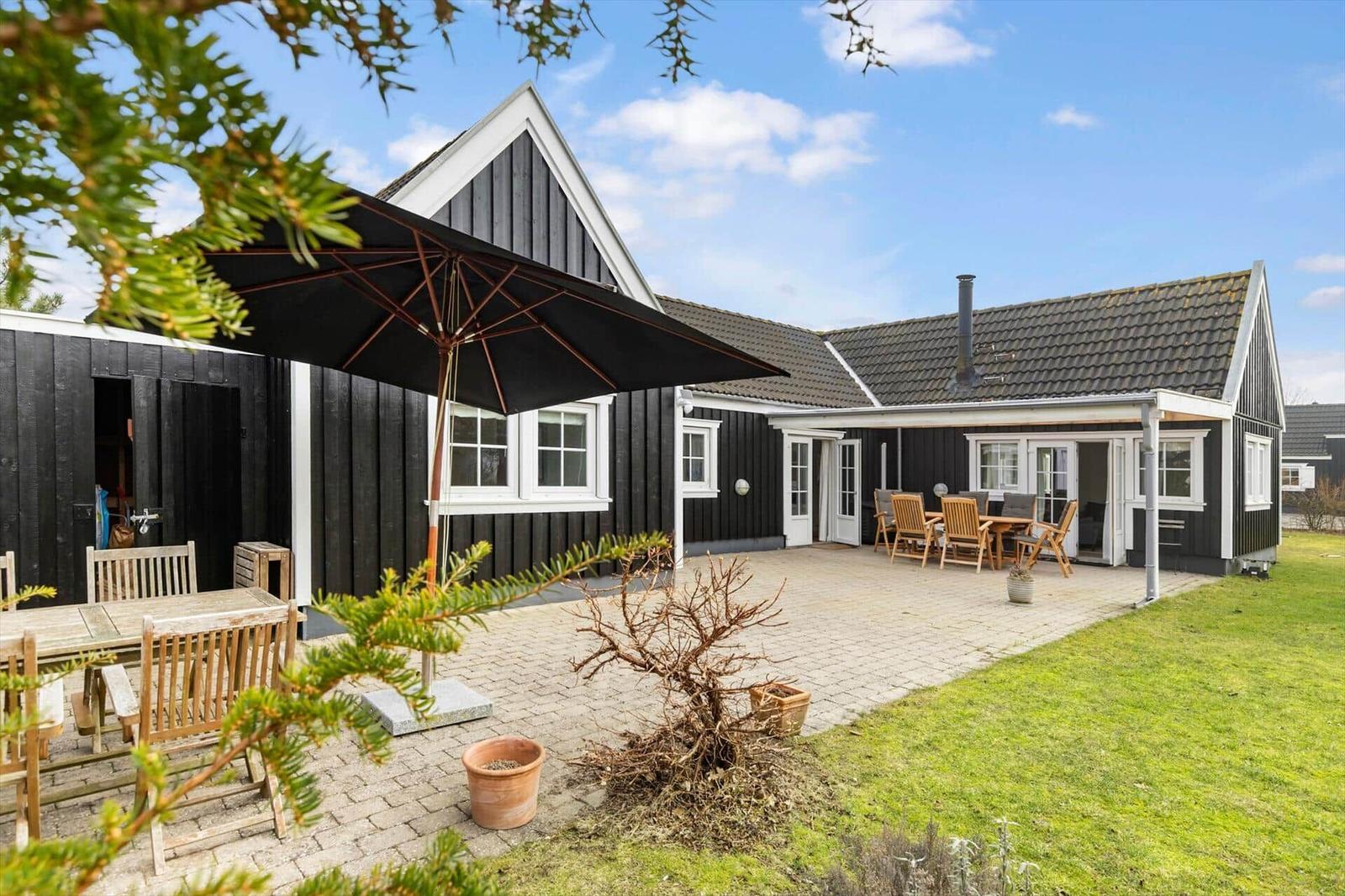 Black wooden house with patio, garden, and umbrella. Wooden tables and chairs.