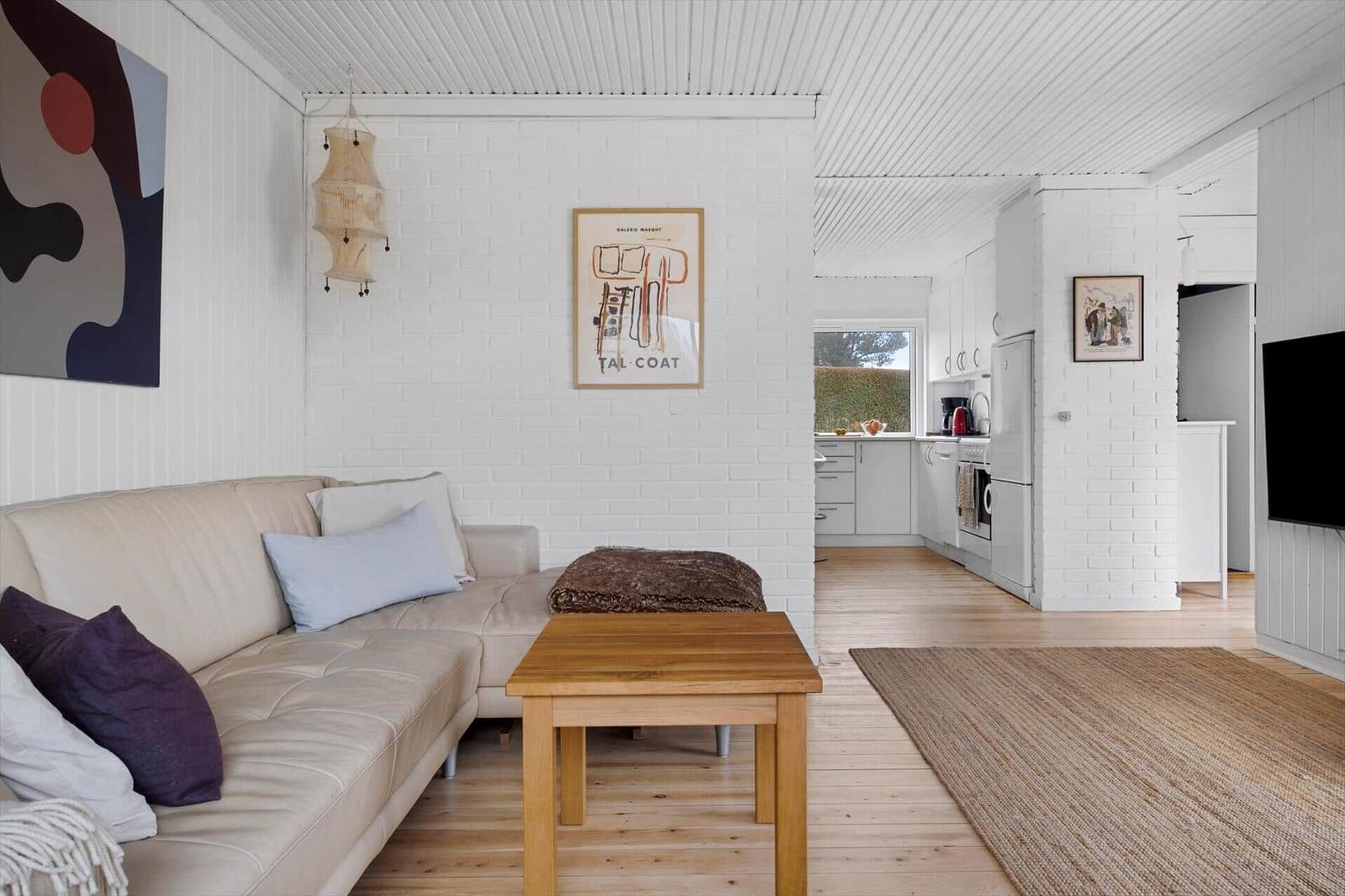 Living room with sofa, table, and kitchen in background. White walls and wooden floor.